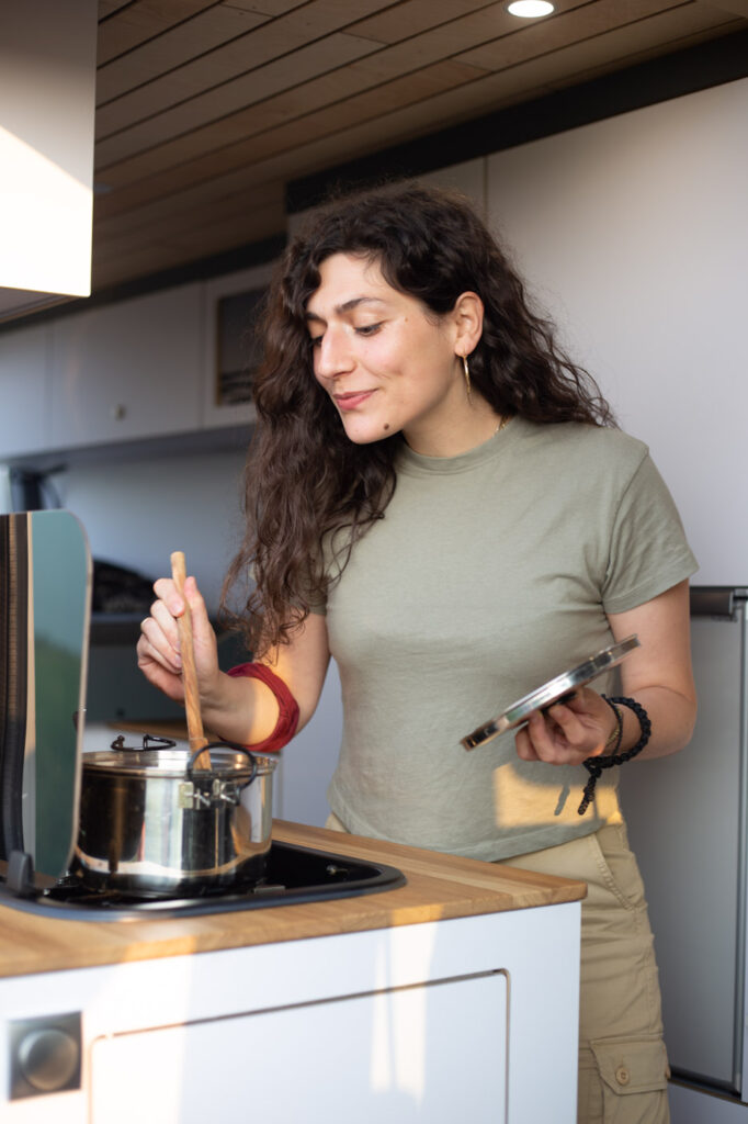 Eine braunhaarige Frau steht an einer Campingküche im von Vanside ausgebauten Wohnmobil, rührt in einem Topf, hält einen Deckel in der Hand und freut sich auf ihr selbstgekochtes Essen in ihrem Campingurlaub.